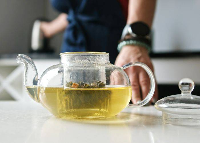 A glass tea pot with tea leaves steeping in the center, turning the hot water a light yellow.